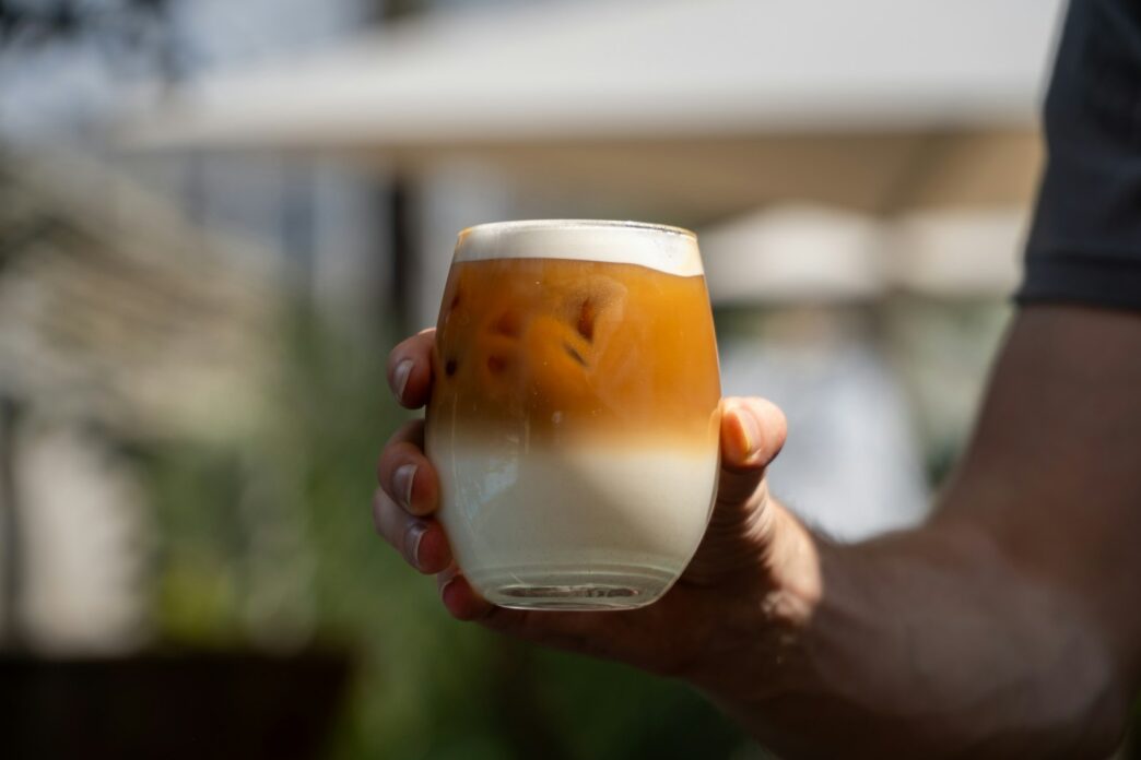 A tall glass of layered iced cloud coffee, with a pale cream float sitting above the darker coffee and coconut water below