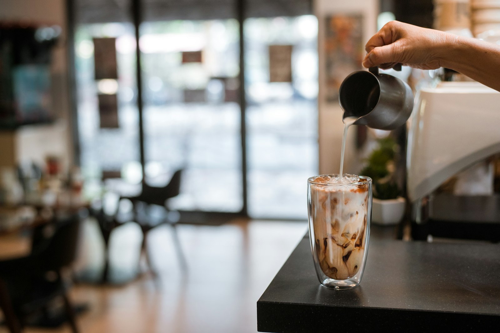 Milk being poured slowly into a glass of iced coffee to form a floating cloud layer
